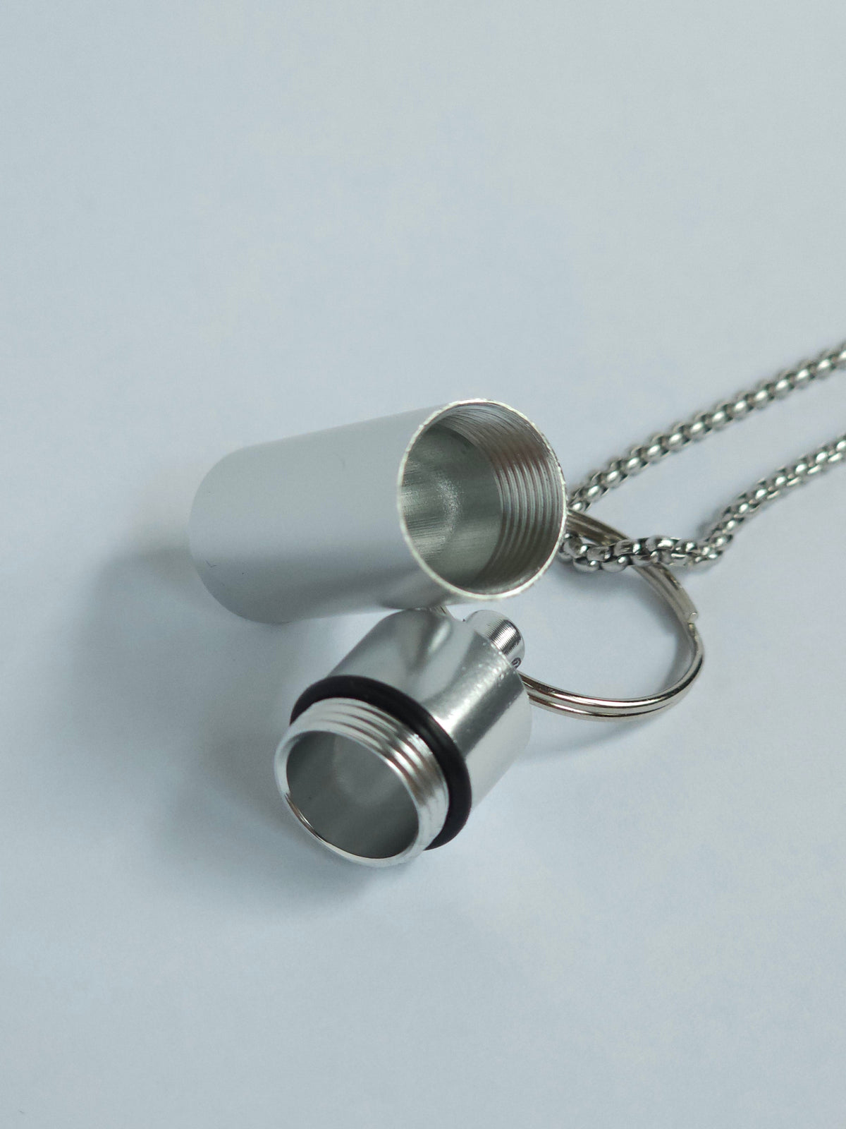 A silver metal pendant discreet snuff storage necklace with a threaded cylindrical design, shown open with its cap removed, attached to a silver ball chain on a light gray background.