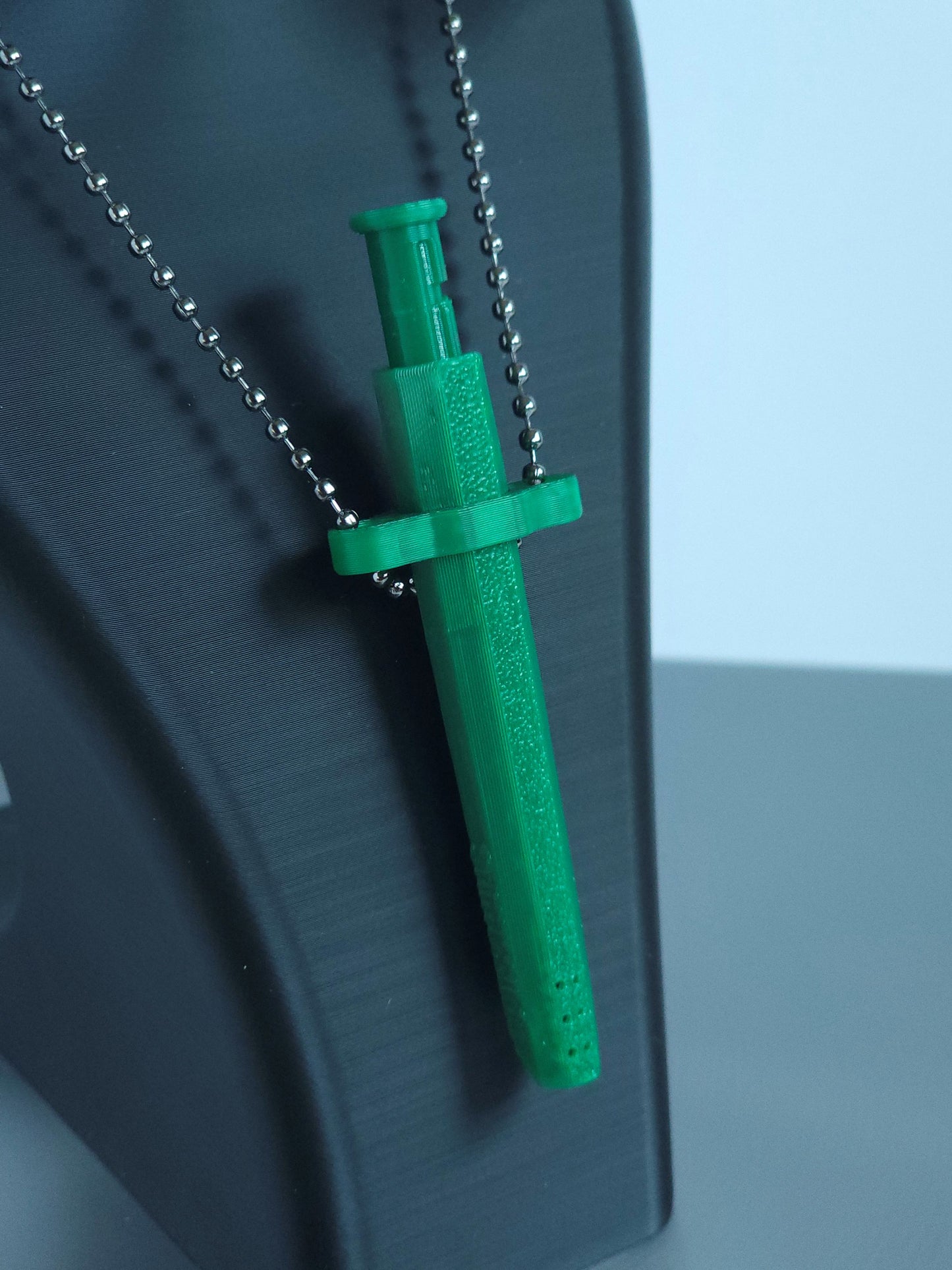 A closeup of a Sniff & Slide spring-loaded dispenser pendant in emerald Green with silver ball chain, displayed on black mannequin bust. The slightly out of focus background is white and grey.